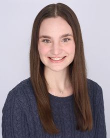 A woman smiling in a blue sweater with a grey background. 