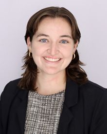 A woman smiling in a black blazer with a grey background.